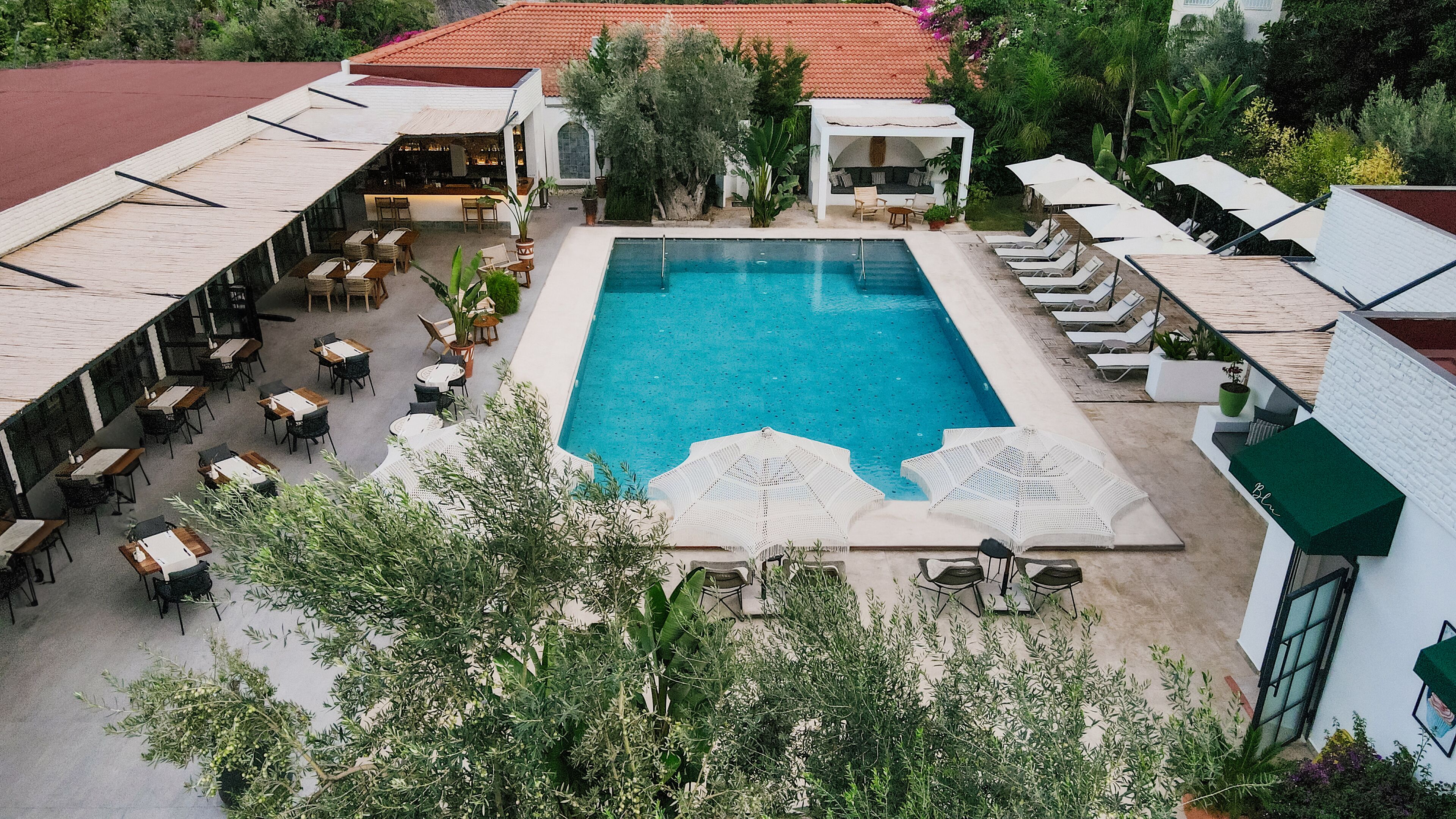 An outdoor swimming pool surrounded by lounge chairs and tables, with a covered seating area and lush greenery in the background.