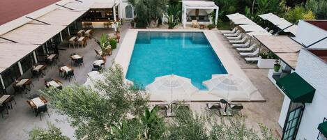 An outdoor swimming pool surrounded by lounge chairs and tables, with a covered seating area and lush greenery in the background.