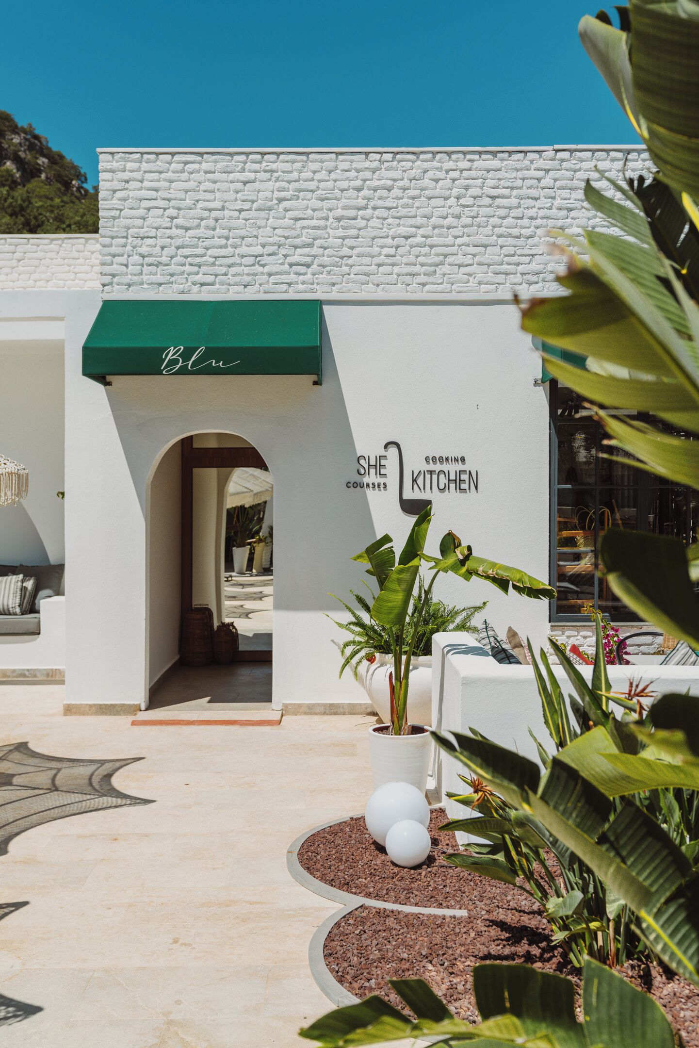 A modern building with a white facade and a green awning. There is a sign that reads "She Kitchen Cooking Courses."