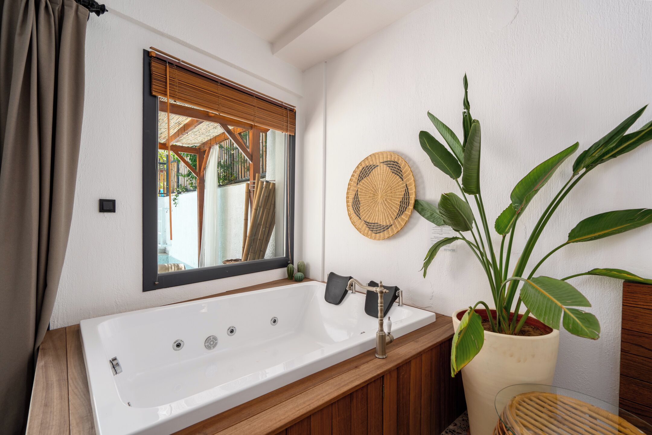 A bathroom with a large bathtub, a window with blinds, a potted plant, and a woven wall hanging.