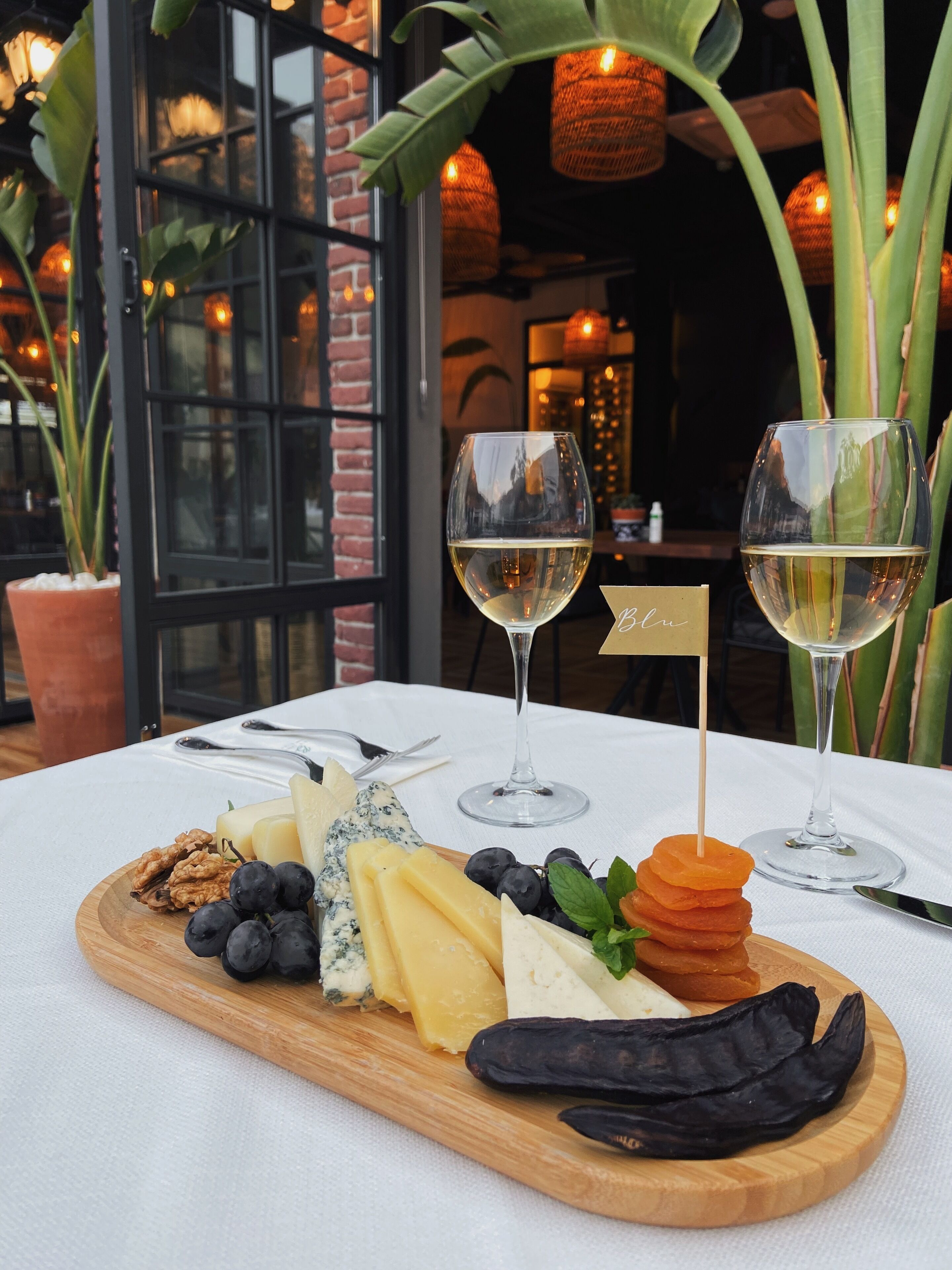 Une planche en bois sur laquelle sont disposés du fromage, des raisins, des noix et des piments séchés. Deux verres de vin blanc sont posés sur la table.