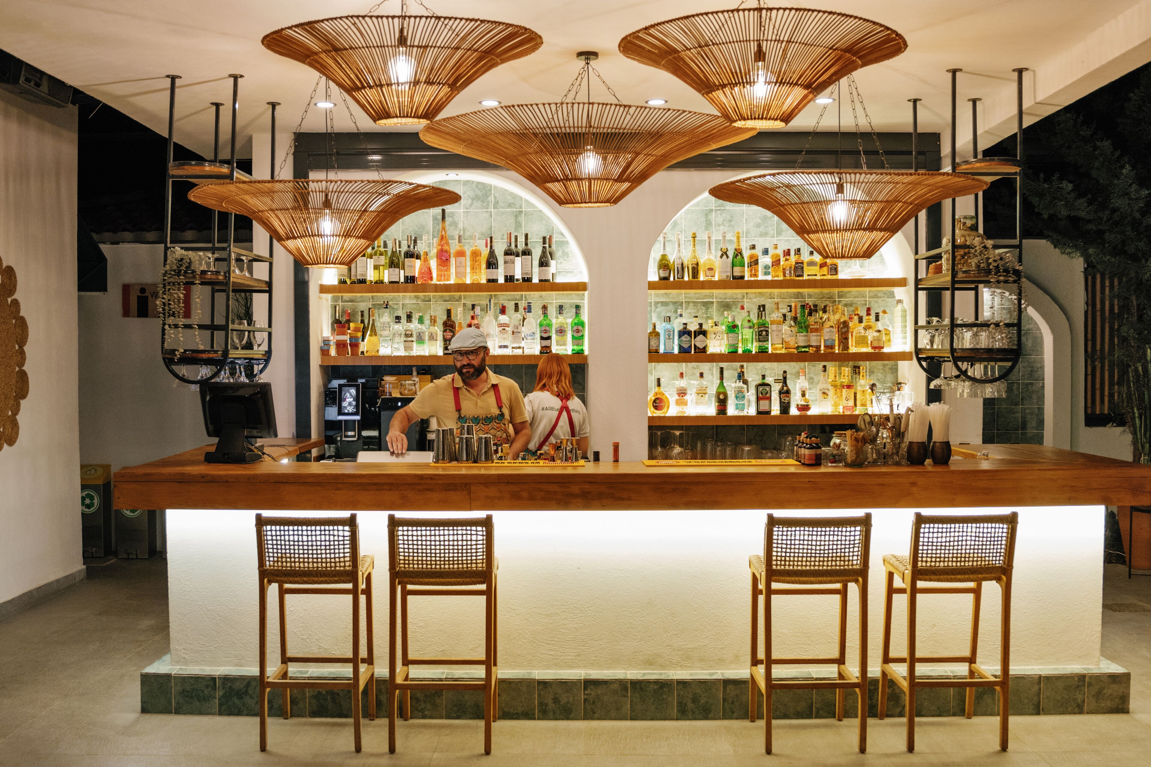 Un bar con mostrador de madera, sillas de mimbre y estantes llenos de botellas.