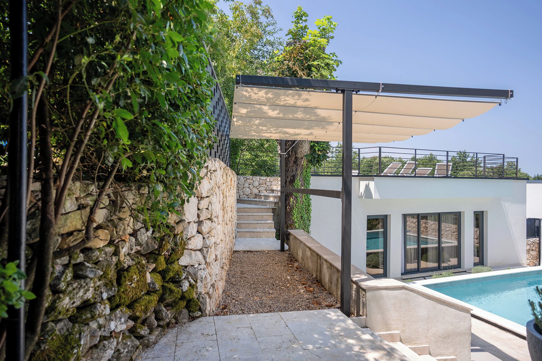 Un área de piscina moderna al aire libre con un muro de piedra, una zona de estar cubierta y vistas despejadas a la piscina.