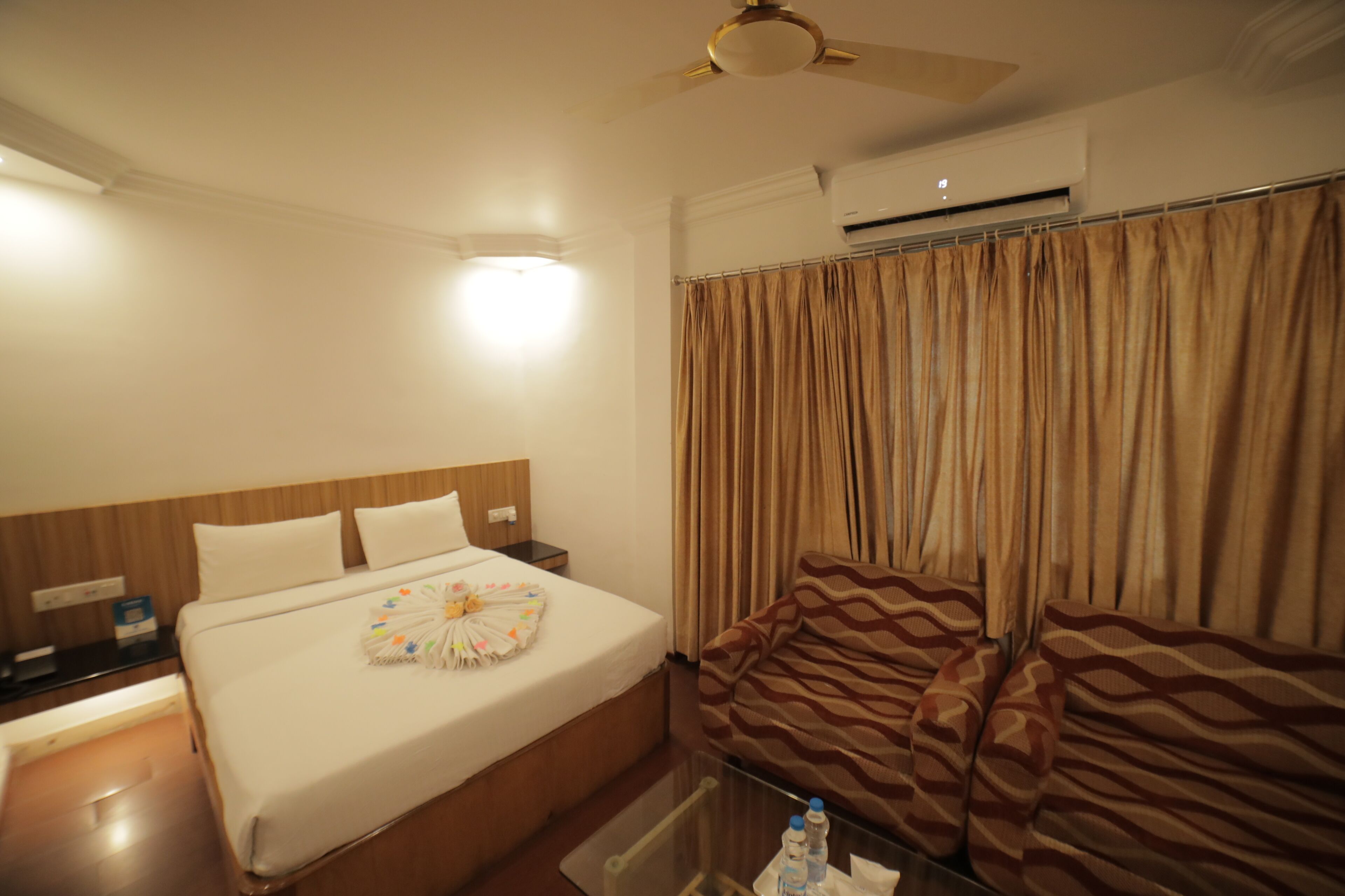 A hotel room with a bed, a nightstand, a chair, a table, and an air conditioning unit.
