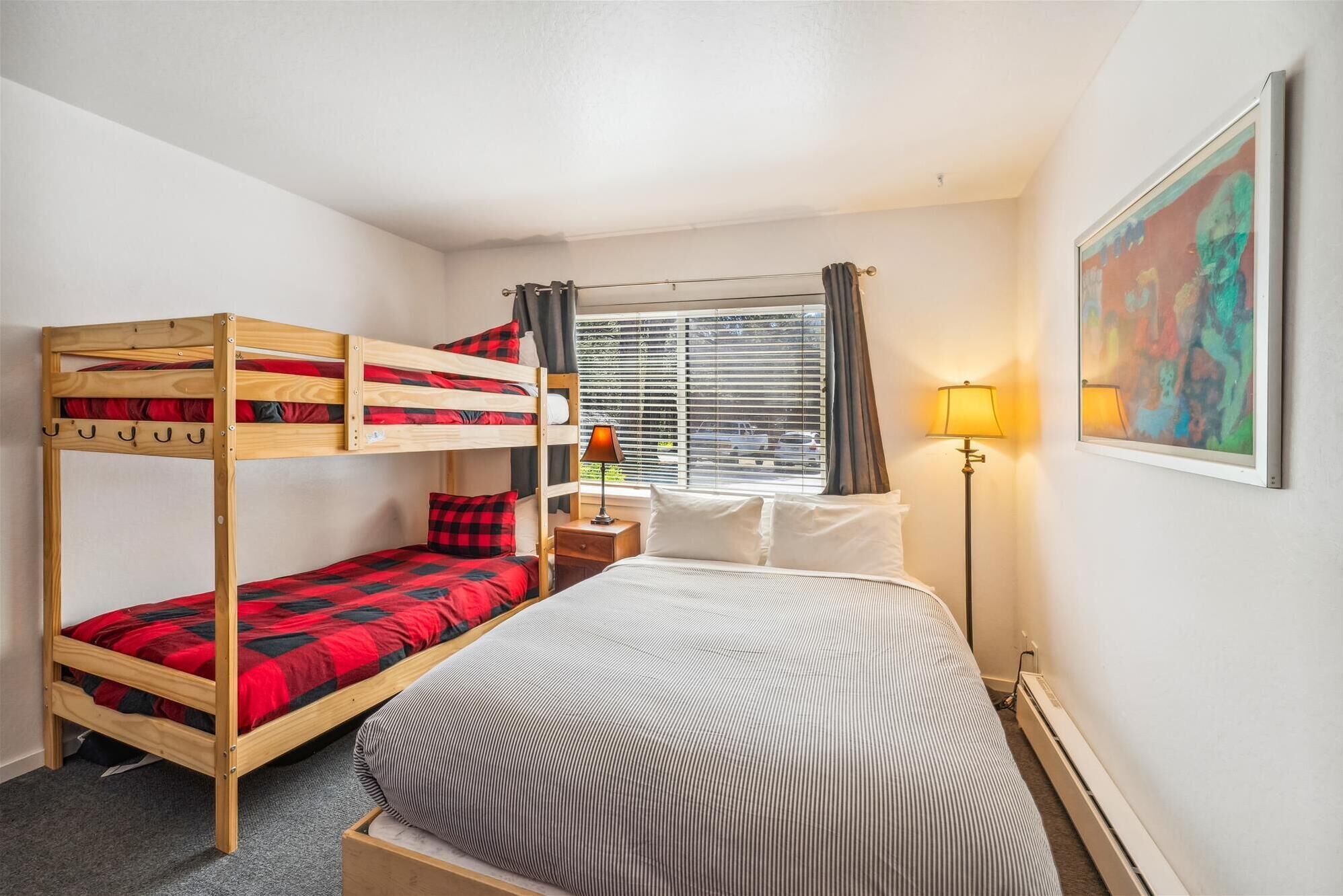 A room with bunk beds, a bedside table with a lamp, a window with curtains, and a painting on the wall.