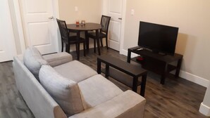 A living room with a sofa, a coffee table, a TV on a stand, and a dining table with chairs. - Heritage 2 Bedroom Deluxe Apartment (Fort McMurray)