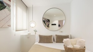 Ein Schlafzimmer mit einem Bett, Kissen, einem runden Spiegel, einer Nachttischlampe und einem Wäschekorb mit Handtüchern.
