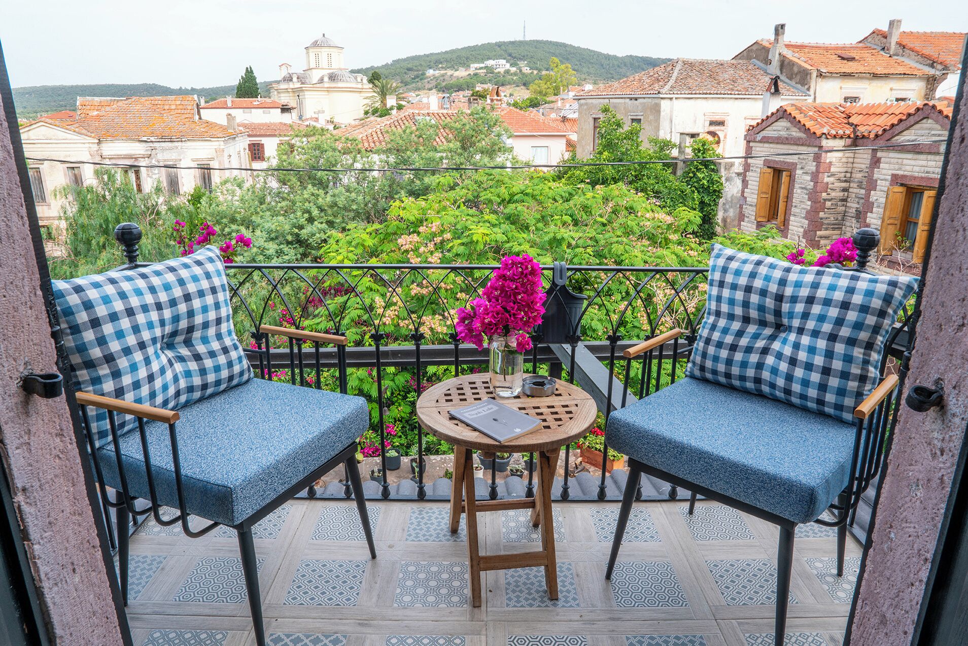 Un balcón con dos sillas azules a cuadros, una mesa redonda pequeña con un libro y un florero con flores rosas. El balcón tiene vistas a un paisaje urbano con edificios y zonas verdes.