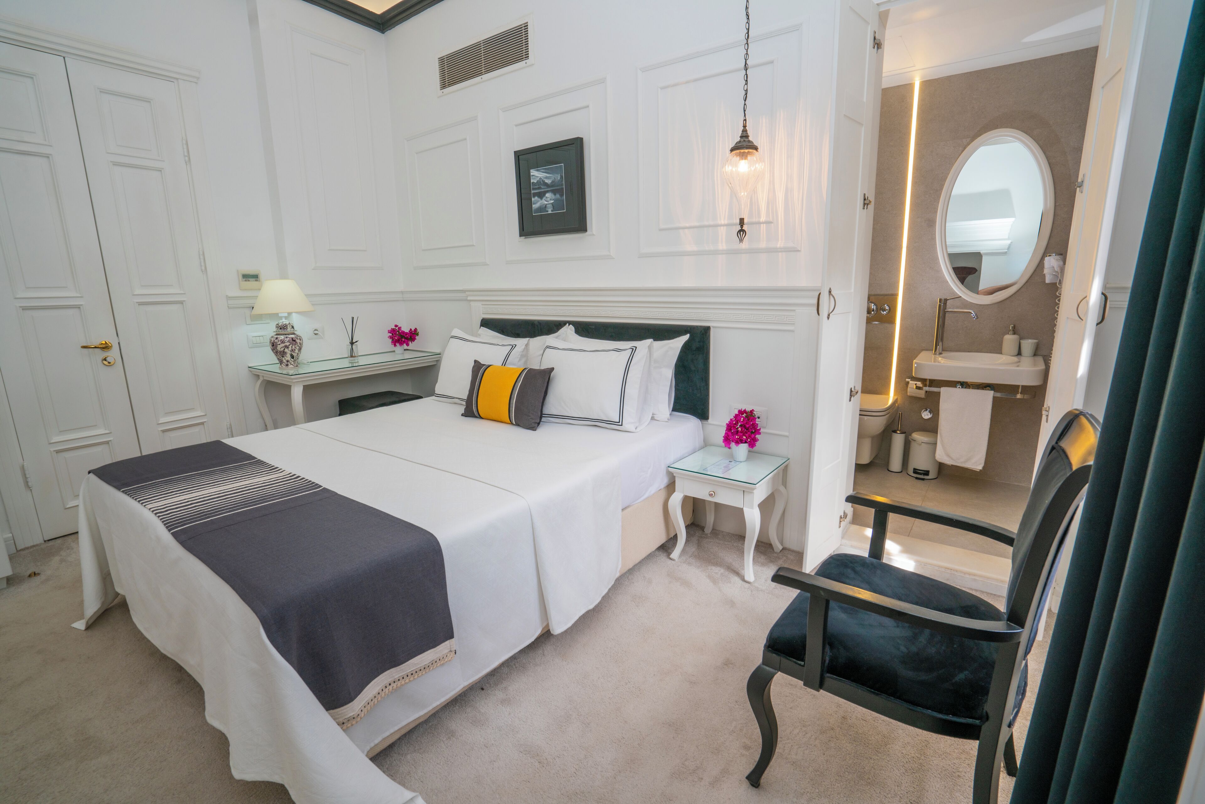 Un dormitorio ordenado con cama, mesitas de noche, una silla y un baño visible a través de una puerta abierta.