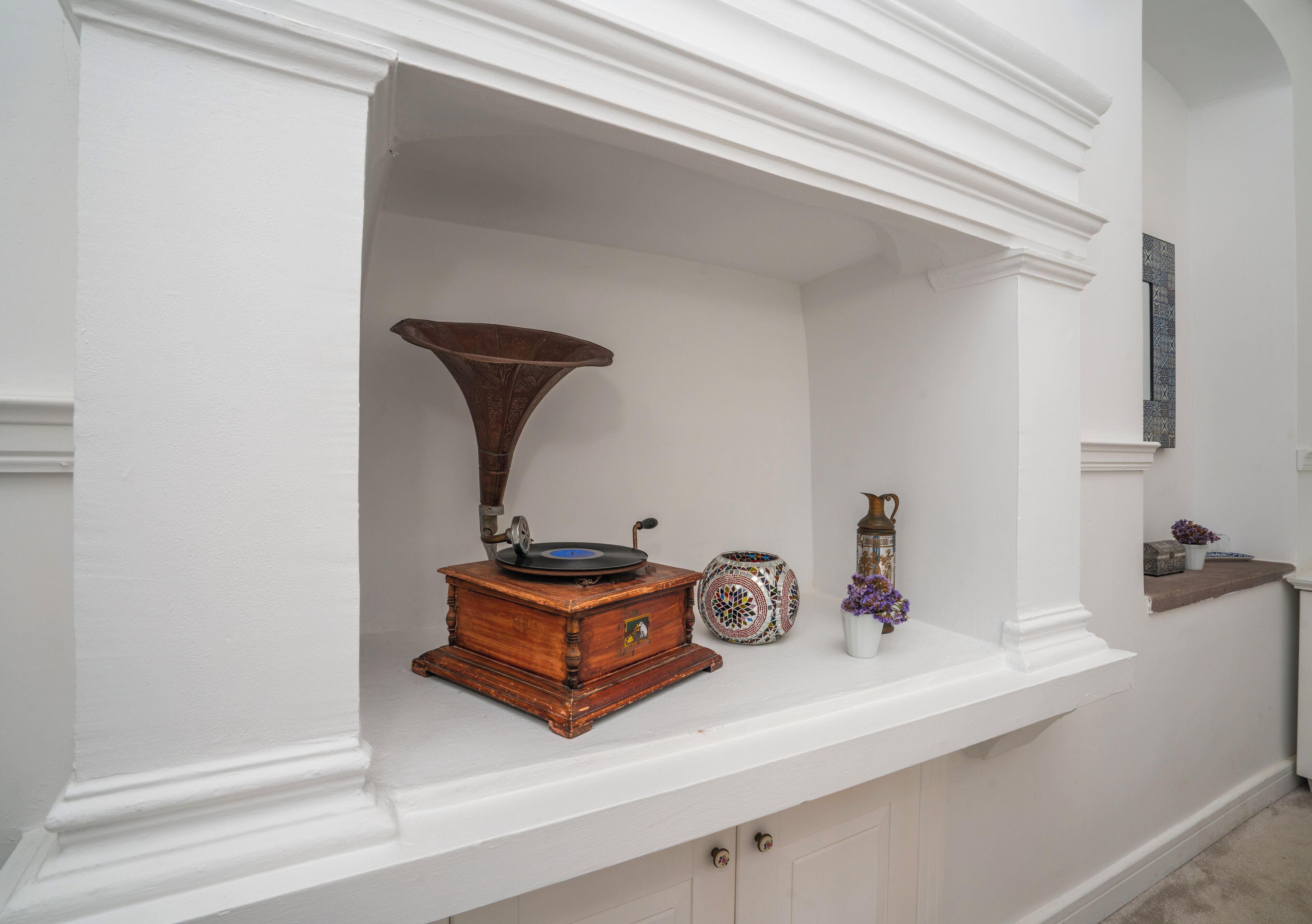 A white wall with a shelf containing a wooden record player, a decorative vase, and a small table with a vase and flowers.