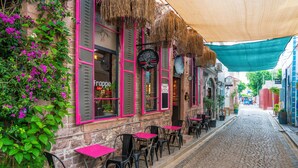 A cobblestone street lined with outdoor seating, a restaurant with pink shutters, and a thatched roof. - Siyah Lale Cunda 1841 (Ayvalik)