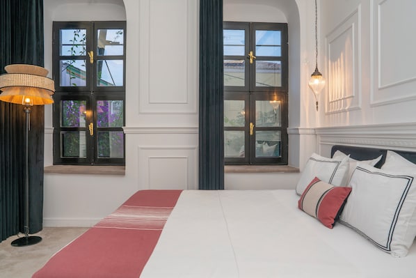 A bedroom with a large bed, two windows with dark frames, a hanging lamp, and a bedside table.
