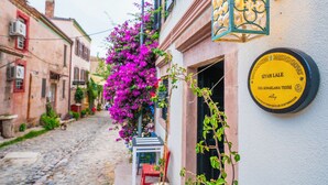 A narrow street with cobblestone pavement, a building with a yellow plaque, and vibrant purple flowers hanging over the entrance. - Siyah Lale Cunda 1841 (Ayvalik)