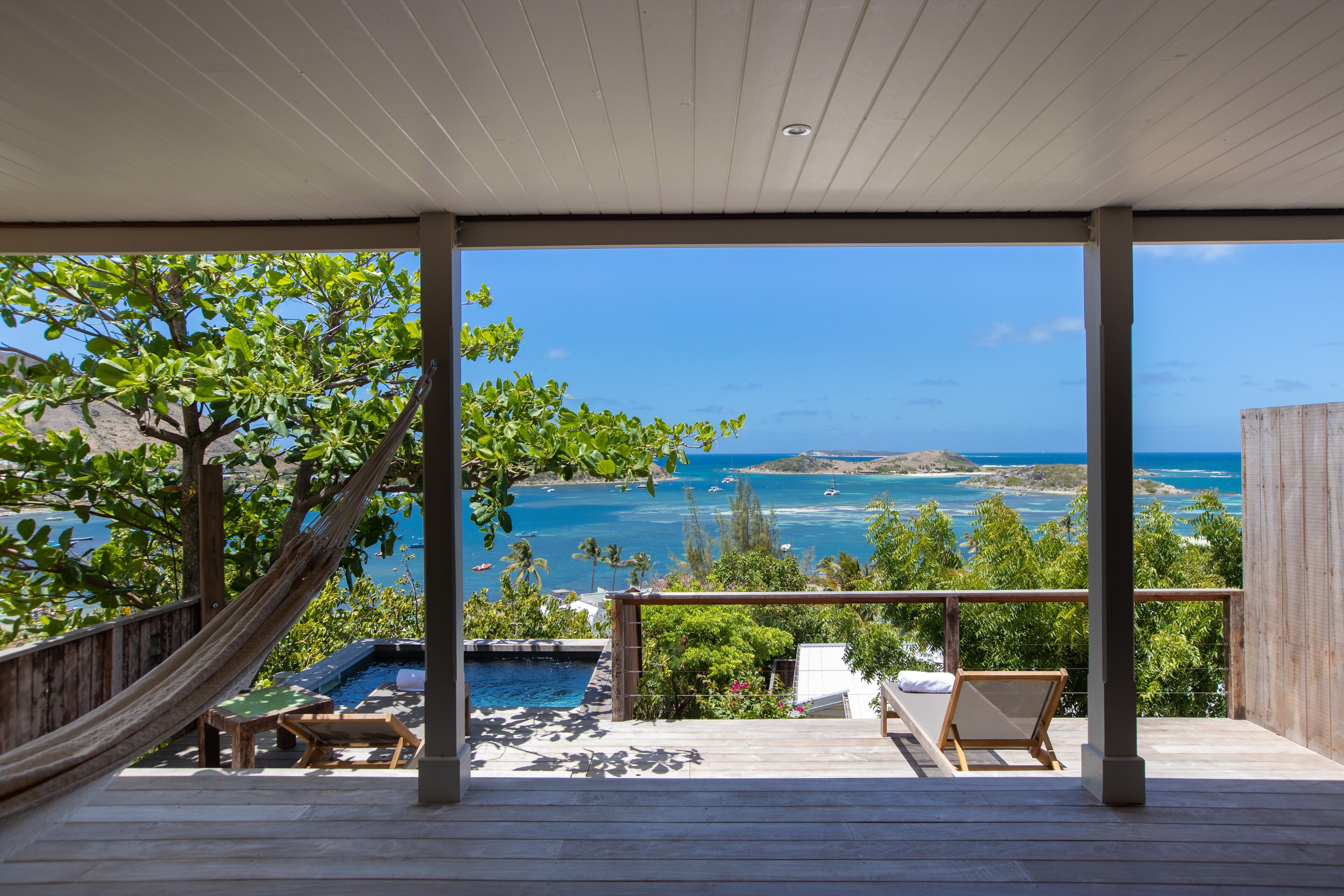 A wooden deck with a hammock, a pool, and a view of the ocean and islands.