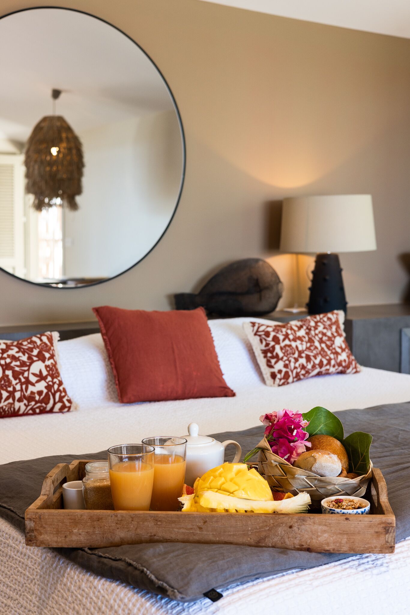 Bandeja de desayuno ordenada con jugo de naranja, un bol de fruta, pan y un recipiente pequeño con nueces sobre una cama.