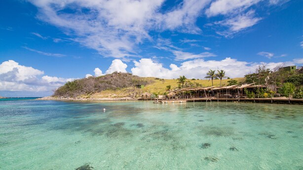 Un paysage côtier avec une eau turquoise et limpide, une plage de sable et une petite colline parsemée de quelques plantes et abritant quelques constructions.