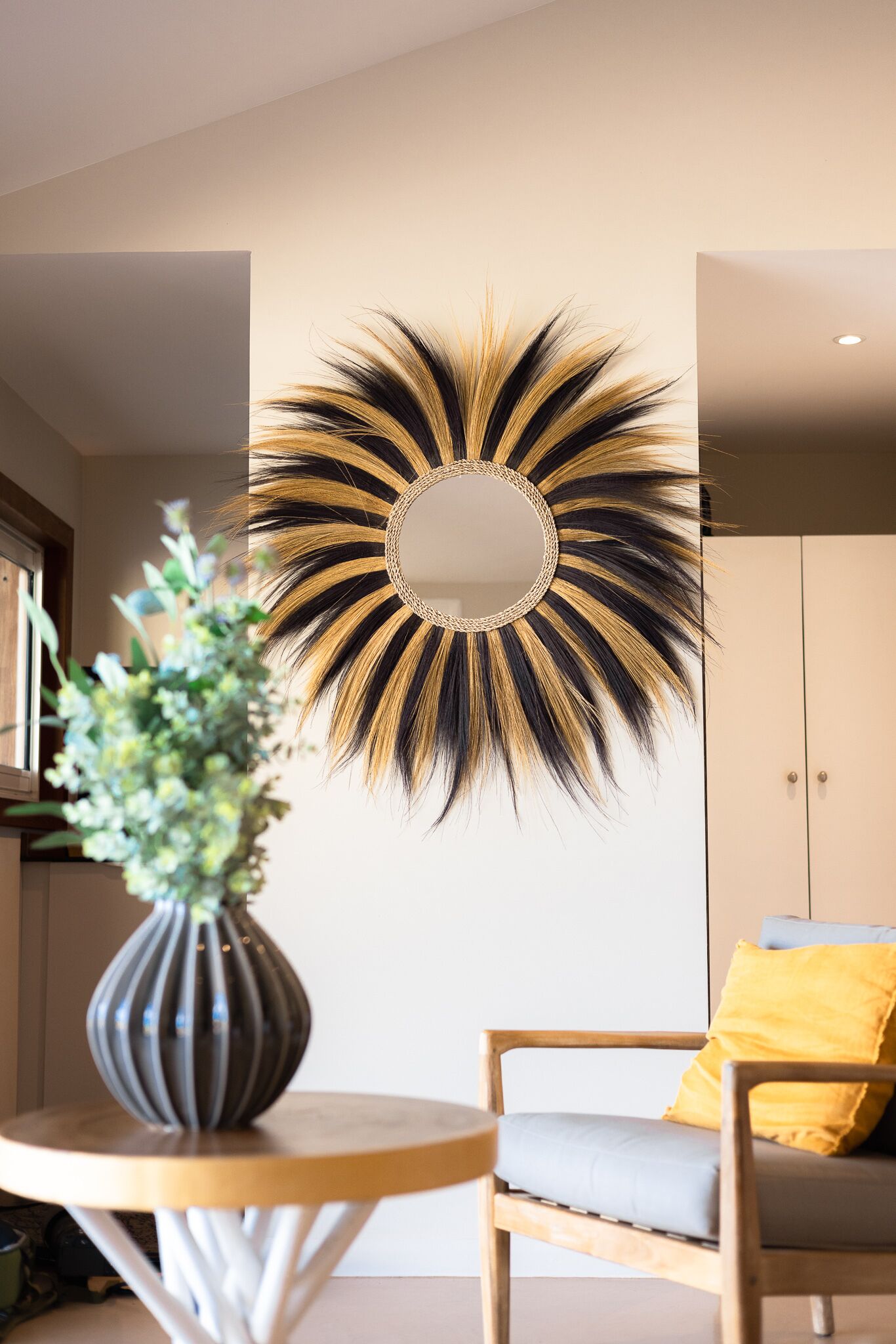 Une pièce avec un grand miroir rond orné d’un motif en forme de soleil sur le mur, une petite table ronde avec un vase, et une chaise avec des coussins.