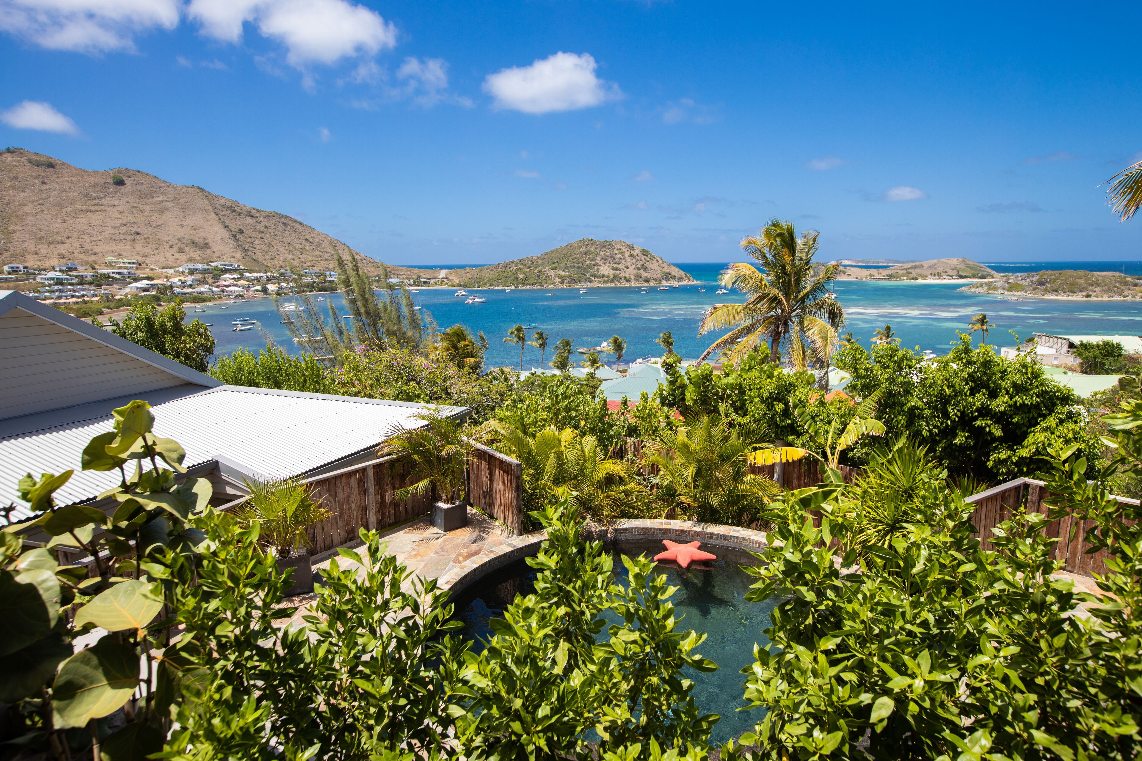 Uma piscina rodeada por vegetação exuberante, com vista para uma área costeira com barcos e colinas.