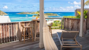 Une terrasse en bois avec un hamac, deux tabourets de bar et un fauteuil inclinable, le tout offrant une vue sur un océan turquoise et des îles à l’horizon.
