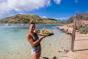 A person holding a tray with food at a beach with clear water and a wooden dock.