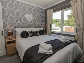 Una cama bien hecha con una decoración en forma de corazón en la pared, una lámpara de noche y vistas del exterior a través de una ventana con cortinas.