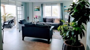 Amplia sala de estar con sofá, mesa de centro y ventilador de techo. Se aprecia un ventanal con cortinas, un cuadro en la pared y una planta en maceta.