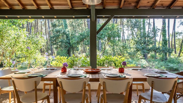 Un área de comedor al aire libre con una mesa de madera preparada para seis personas, rodeada de sillas, bajo un pérgola de madera con vista a una zona boscosa.