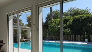 Area piscina interna moderna con porte in vetro che si affacciano su una piscina esterna, immersa nel verde e delimitata da una recinzione in legno.