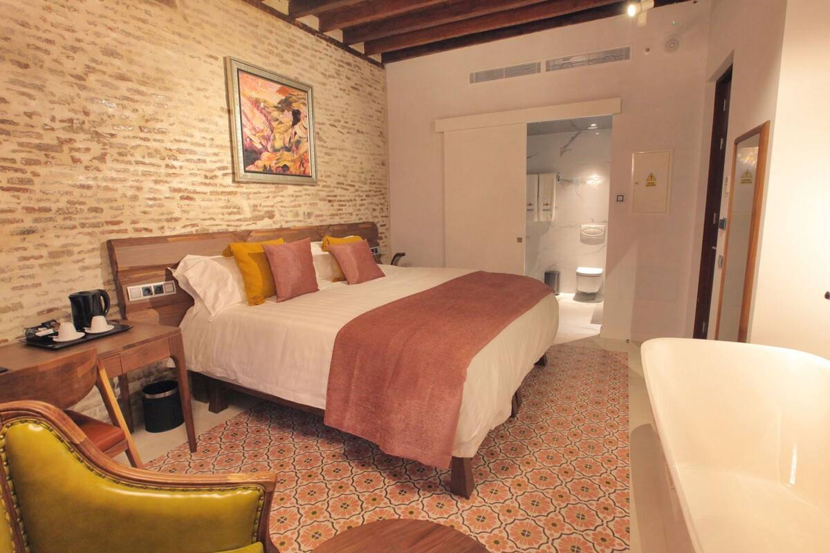 Una habitación de hotel con pared de ladrillo, una cama con manta rosa, una mesita de noche de madera, una silla amarilla, una bañera blanca y un baño visible a través de una puerta abierta.