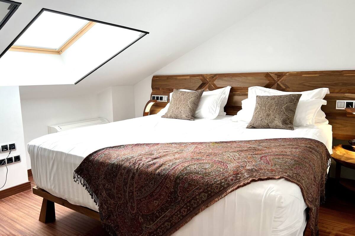 Un dormitorio con tragaluz, cabecera de madera, colcha estampada y mesita de noche con lámpara.