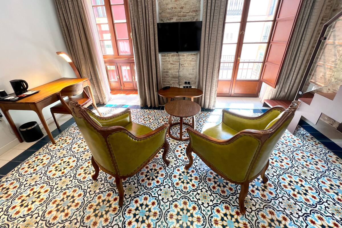 Una habitación con piso de baldosas, dos sillones verdes, una mesita de madera, un televisor montado en una pared de ladrillo y vistas a través de una ventana con cortinas.