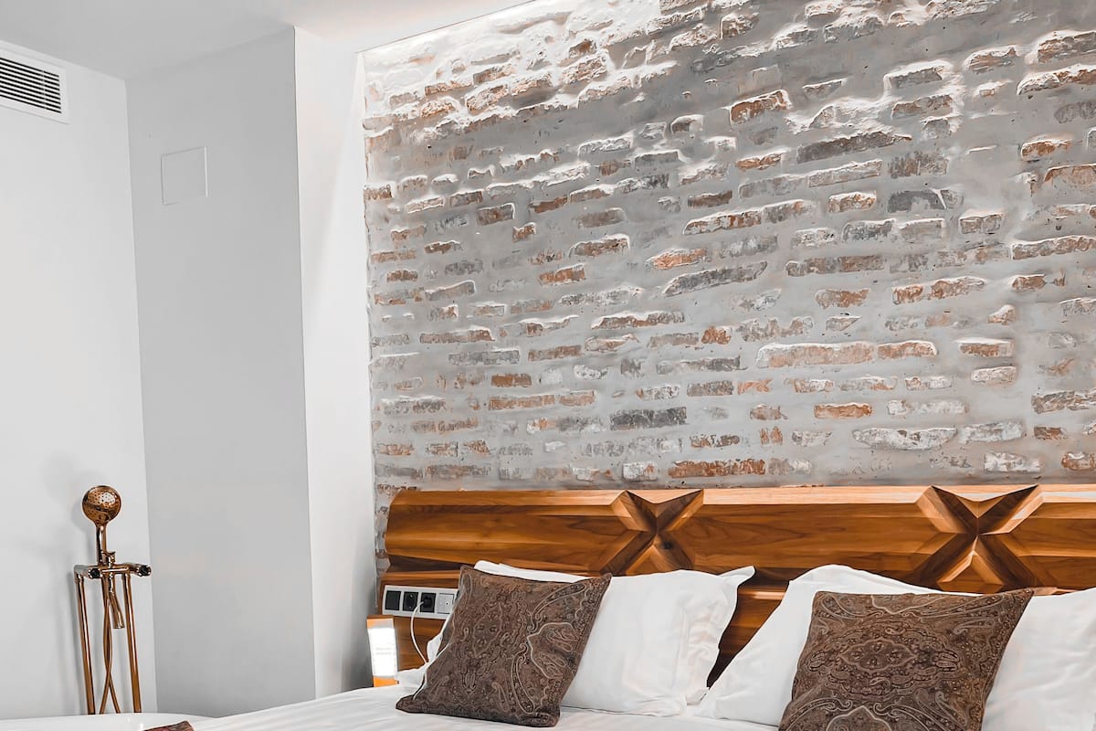 Una cama bien hecha con sábanas blancas y dos almohadas estampadas, un cabecero de madera y una pared de ladrillo detrás.