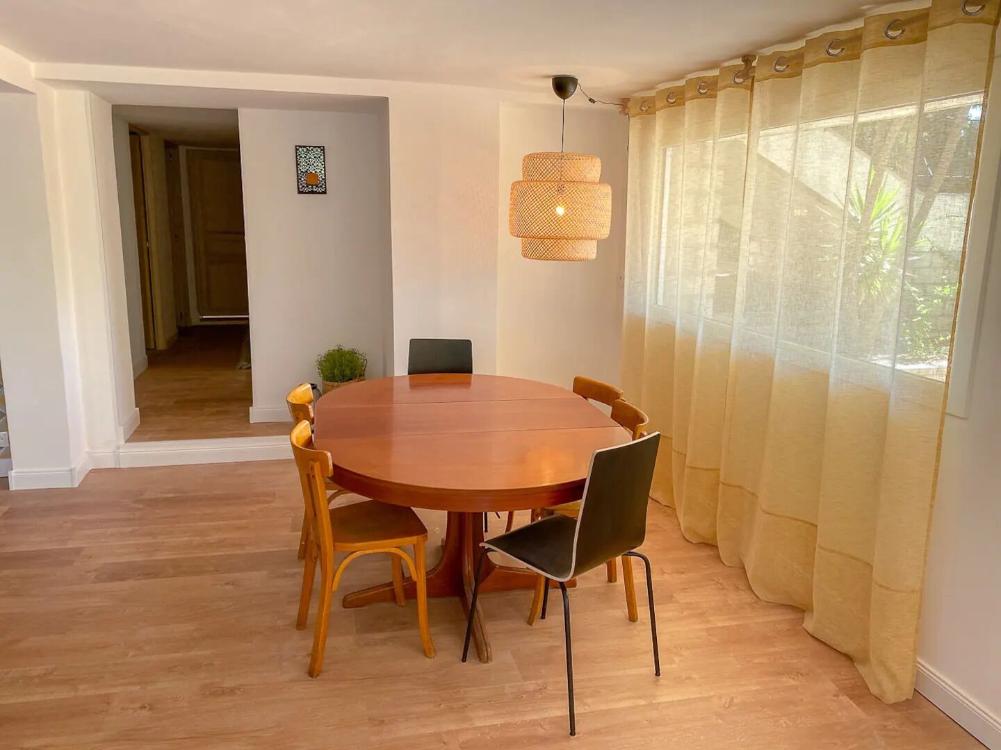 Un coin repas avec une table ronde en bois, six chaises, un luminaire suspendu et une vue dégagée à travers des rideaux légers.