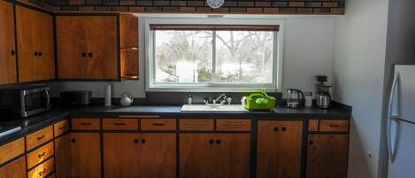 A kitchen with wooden cabinets, a microwave, a sink, and a window with a view of trees.
