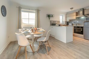 A modern kitchen with white cabinetry, a central island, and a dining area with white chairs and a round table. - 2 Bed in Woolacombe (oc-beache) (Woolacombe)