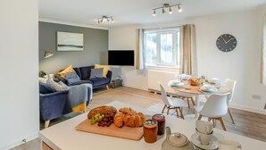 A modern living room with a sofa, a dining table set for breakfast, and a kitchen area with a cutting board and food items.