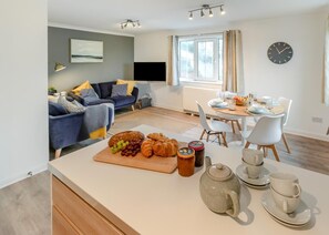 A modern living room with a sofa, a dining table set for breakfast, and a kitchen area with a cutting board and food items. - 2 Bed in Woolacombe (oc-beache) (Woolacombe)