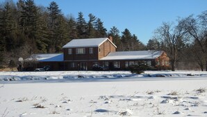 A two-story wooden house with a snow-covered roof, surrounded by a snowy landscape and dense trees.