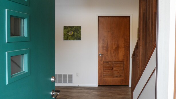 A hallway with a teal door, a wooden door, a small picture on the wall, and a black and blue striped rug.