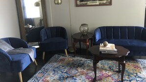 Un salón con dos sillones azules, una mesita de centro, una lámpara de pie, un cuadro con una vista urbana enmarcada y un espejo.