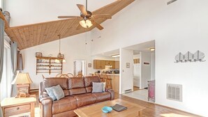 A spacious living room with a brown leather sofa, wooden coffee table, and a ceiling fan. A kitchen is visible in the background. - Birds Nest- Hot Tub w/ Mtn View, Whirlpool Bath, Gas Grill, Keyless Entry (Seven Devils)