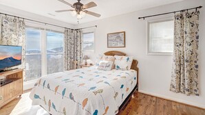 A bedroom with a large bed, a ceiling fan, a TV, and a view of the mountains through the windows. - Birds Nest- Hot Tub w/ Mtn View, Whirlpool Bath, Gas Grill, Keyless Entry (Seven Devils)