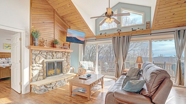 A cozy living room with a stone fireplace, wooden ceiling, and a large window overlooking a deck. - Birds Nest- Hot Tub w/ Mtn View, Whirlpool Bath, Gas Grill, Keyless Entry (Seven Devils)
