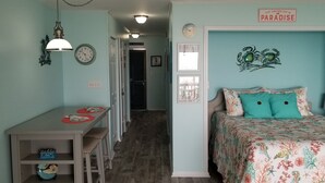 A room with a bed, a desk, and a chair. There is a clock on the wall and a sign that says "Just another day in paradise". - Sea-breezin' - Beachfront Condo                   (Surfside Beach)
