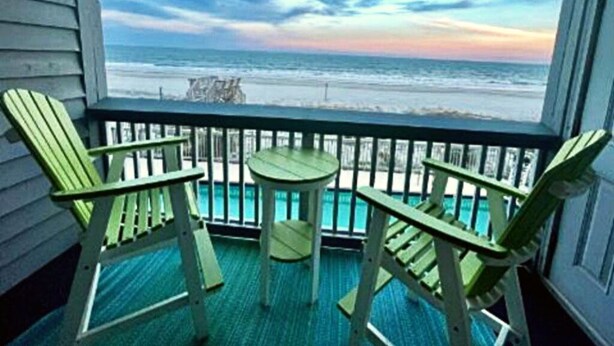 A balcony with two green Adirondack chairs and a small table, overlooking a beach with waves and a sunset sky. - Sea-breezin' - Beachfront Condo                   (Surfside Beach)