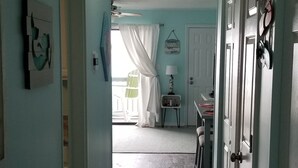 A hallway with light blue walls, wooden flooring, and a ceiling fan. There is a door on the right and a window with curtains in the background. - Sea-breezin' - Beachfront Condo                   (Surfside Beach)