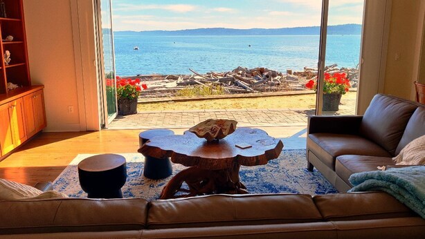 A living room with a large sliding glass door leading to a patio with a view of the sea and a pile of driftwood. - Beach house no-bank waterfront 4-bedroom vacation home on Camano Island (Camano)
