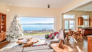 A spacious living room with a large window overlooking a body of water, a wooden dining area, and a kitchen with wooden cabinets. - Beach house no-bank waterfront 4-bedroom vacation home on Camano Island (Camano)