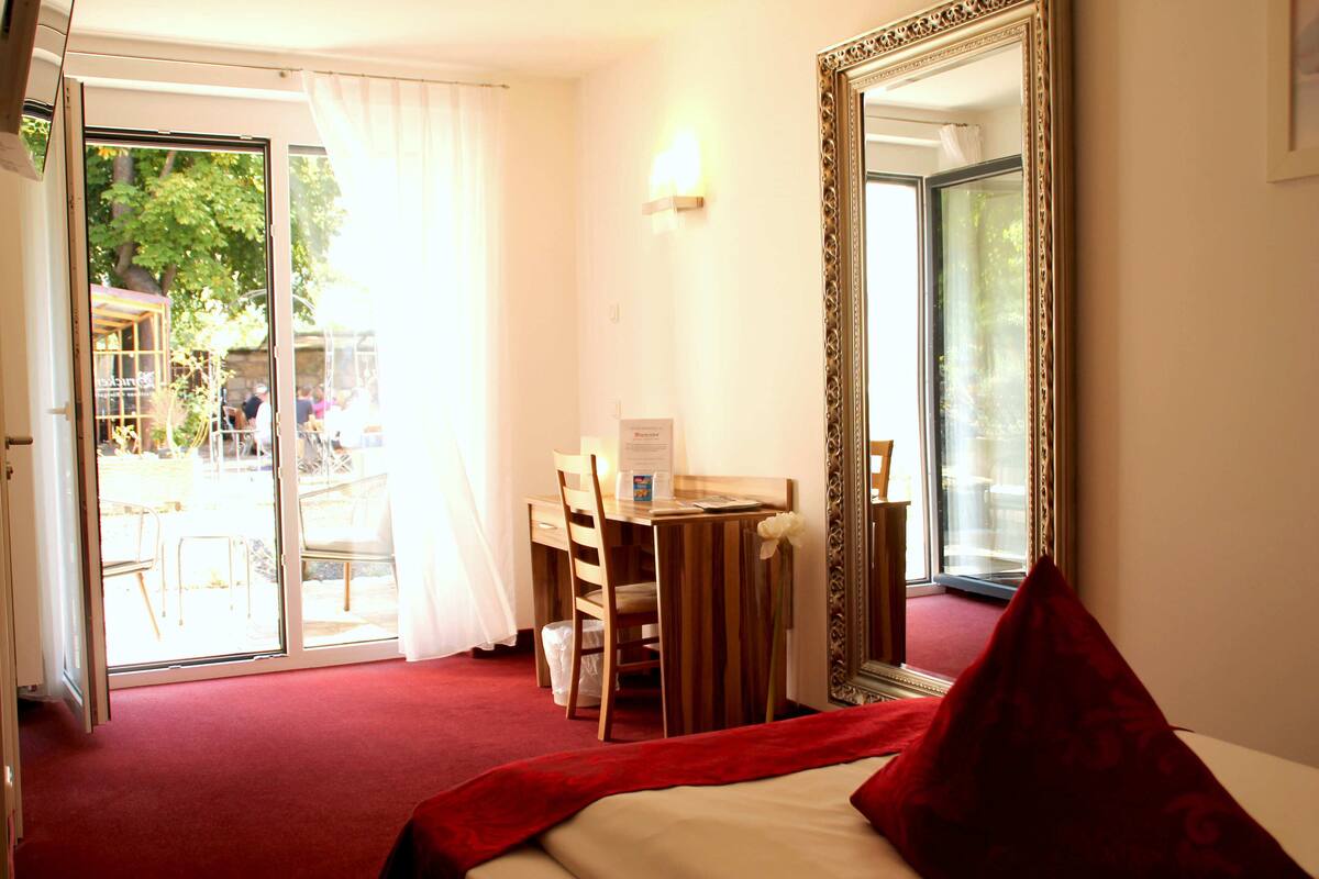 A hotel room with a bed, a desk, and a chair. There is a large mirror on the wall and a sliding glass door leading to an outdoor area.