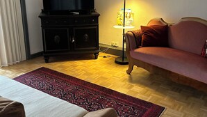 En stue med sofa, fjernsyn på et skab, gulvlampe og et mønstret tæppe.